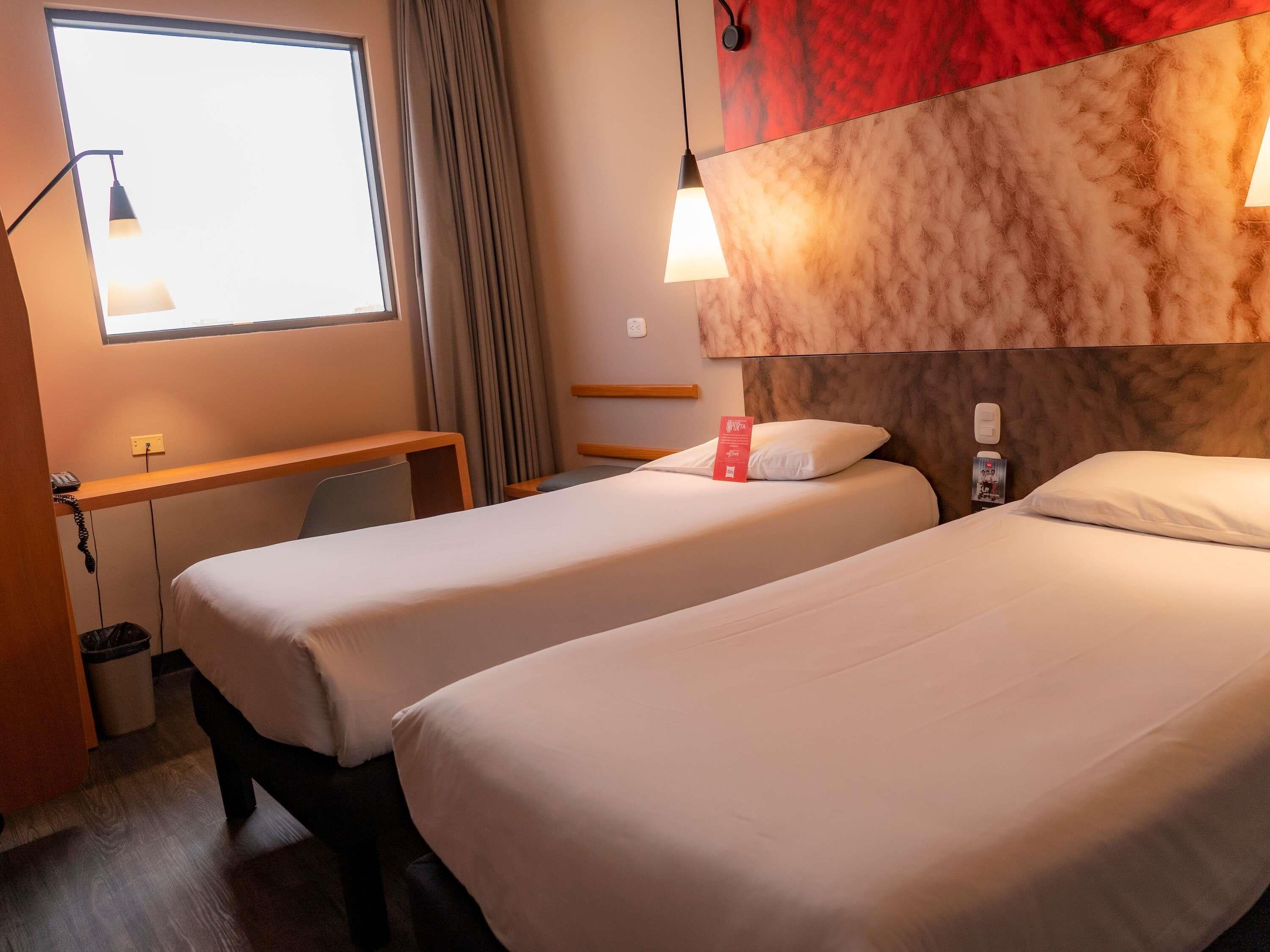 A hotel room with two beds, a desk, a chair, a window with curtains, and a wall with a textured design.