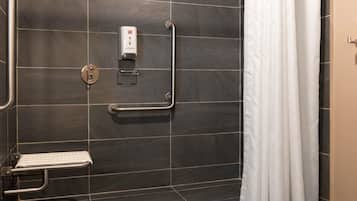 A bathroom with a shower, a hand sanitizer dispenser, a grab bar, and a toilet with a safety bar.