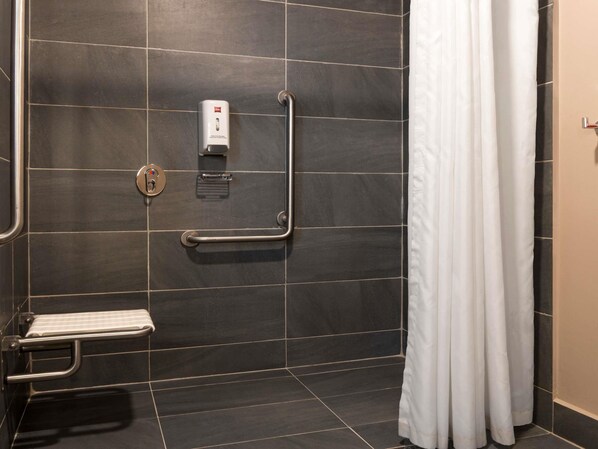 A bathroom with a shower, a hand sanitizer dispenser, a grab bar, and a toilet with a safety bar.