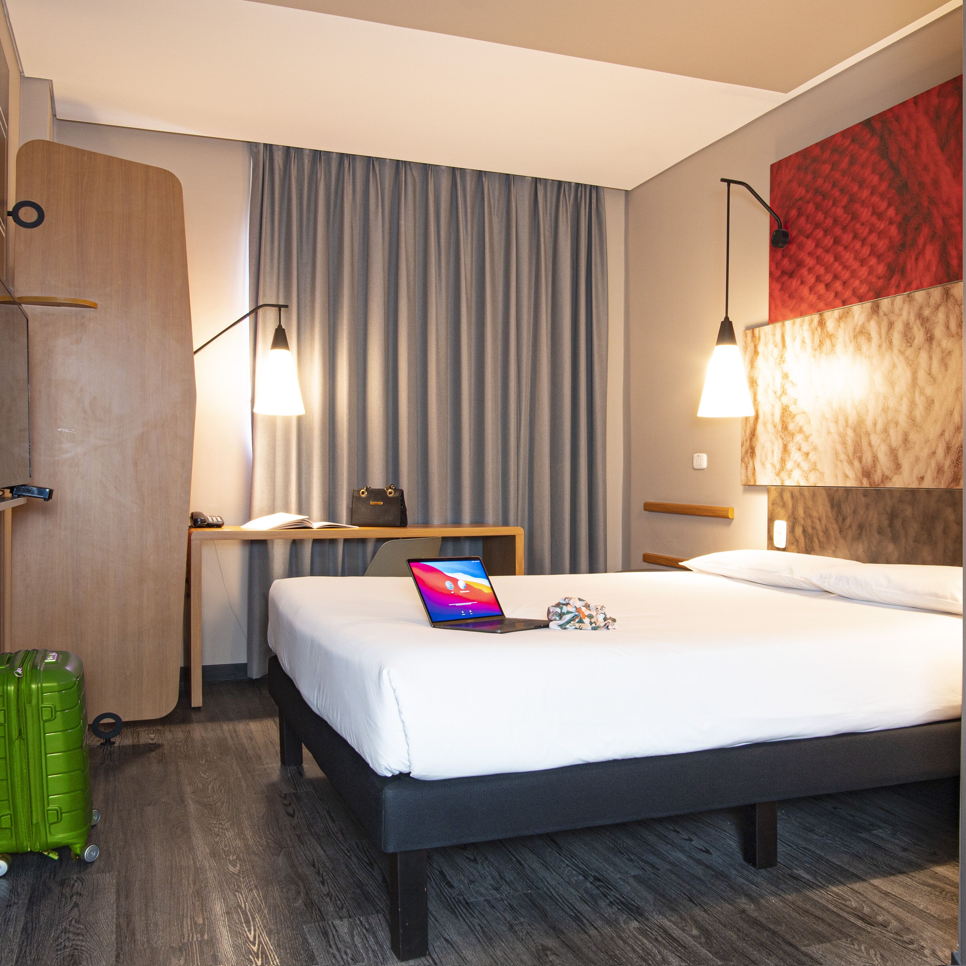 A hotel room with a bed, a desk with a laptop, a suitcase, and a lamp.