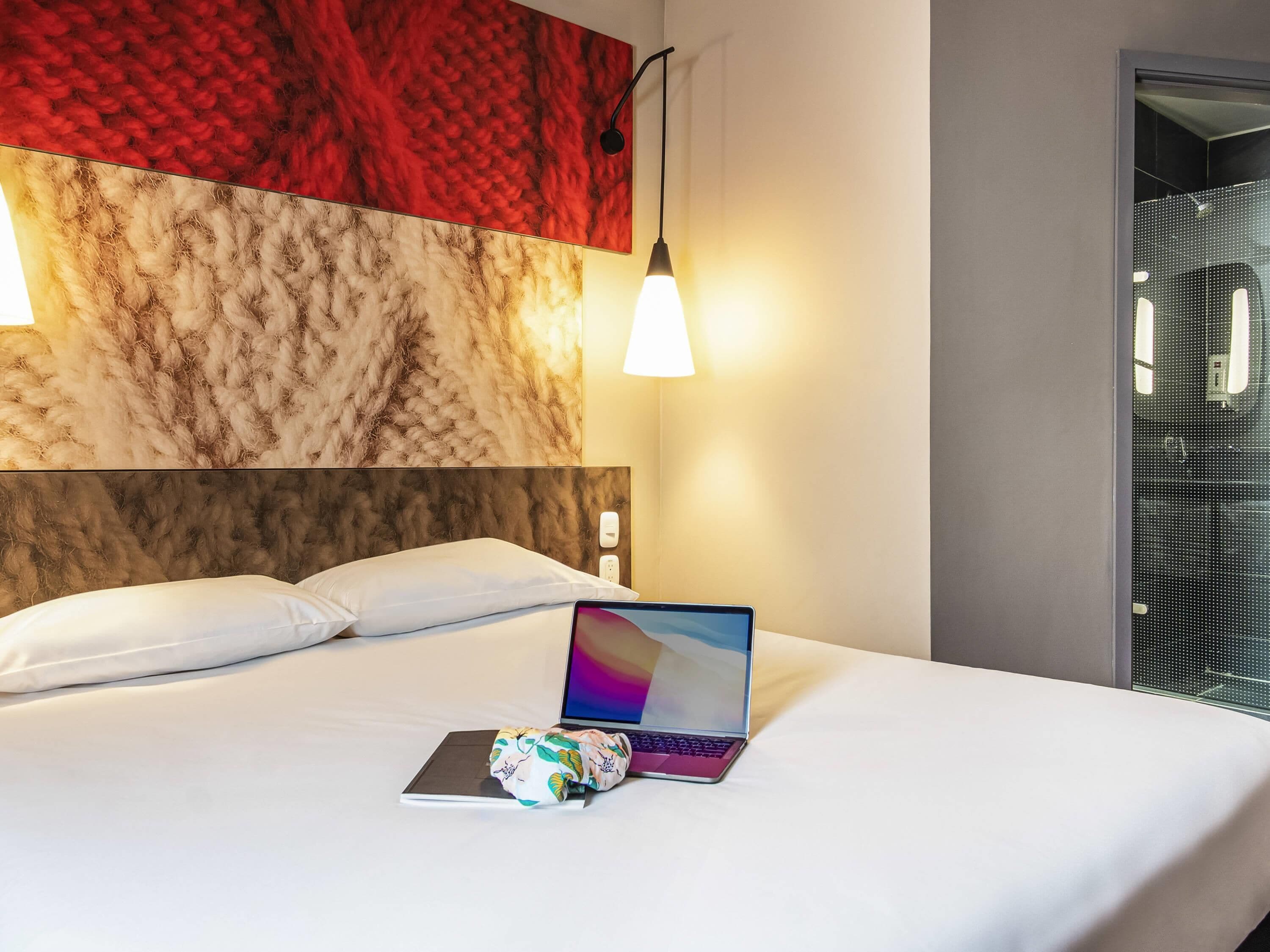 Habitación de hotel con cama, laptop, almohada, pared con textura y una lámpara colgante.