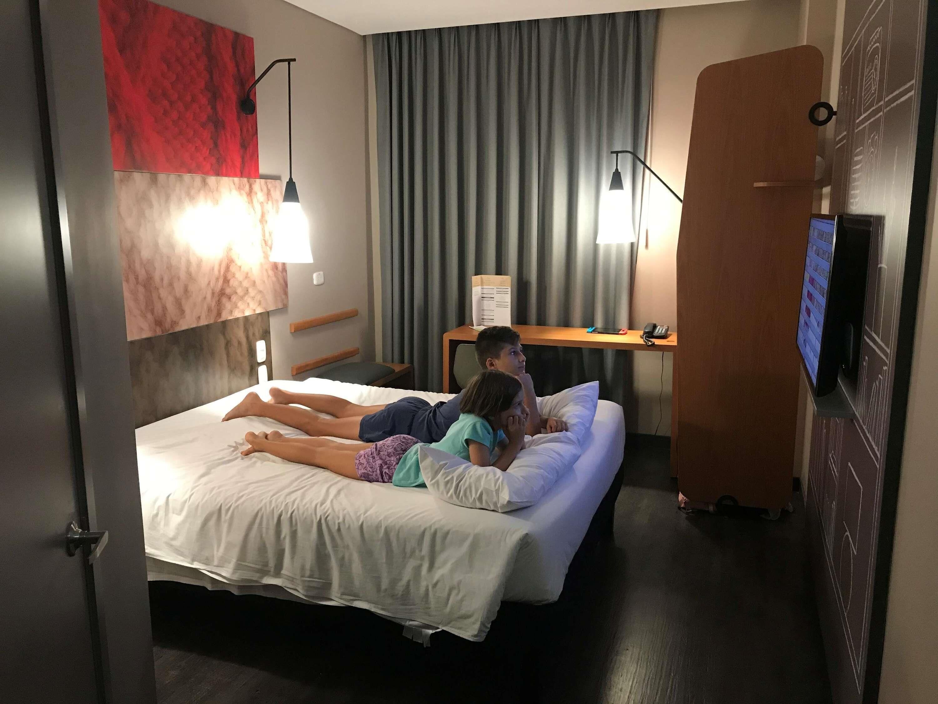 Habitación de hotel con una cama, dos personas acostadas sobre ella, un escritorio con una computadora y un televisor.