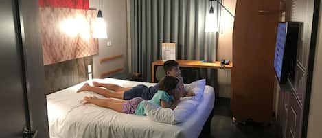 Une chambre d’hôtel avec un lit, deux personnes allongées dessus, un bureau avec un ordinateur et une télévision.