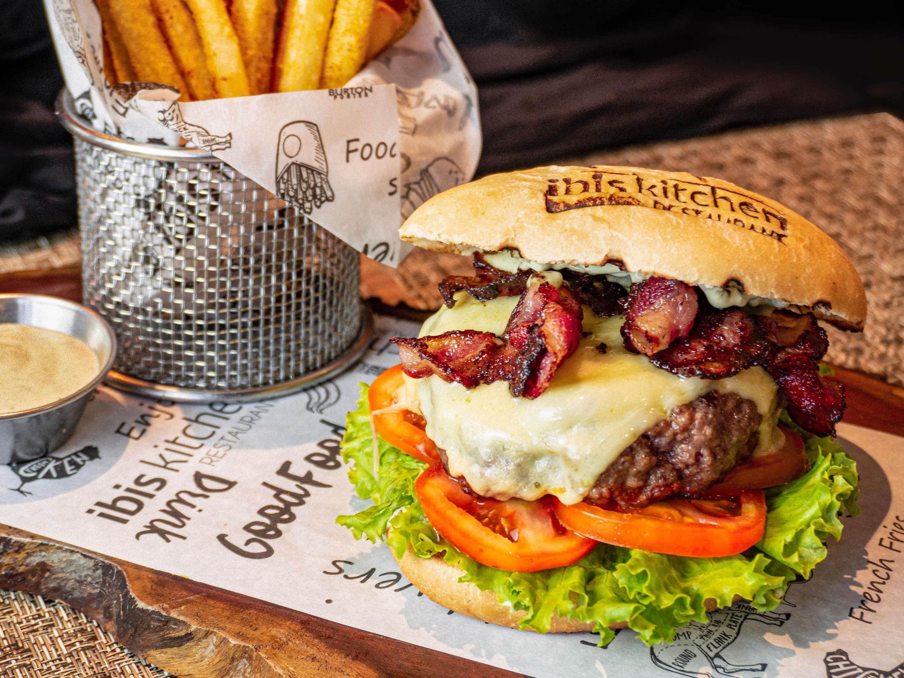 A burger with bacon, cheese, and lettuce on a bun, accompanied by fries and a small container of sauce.