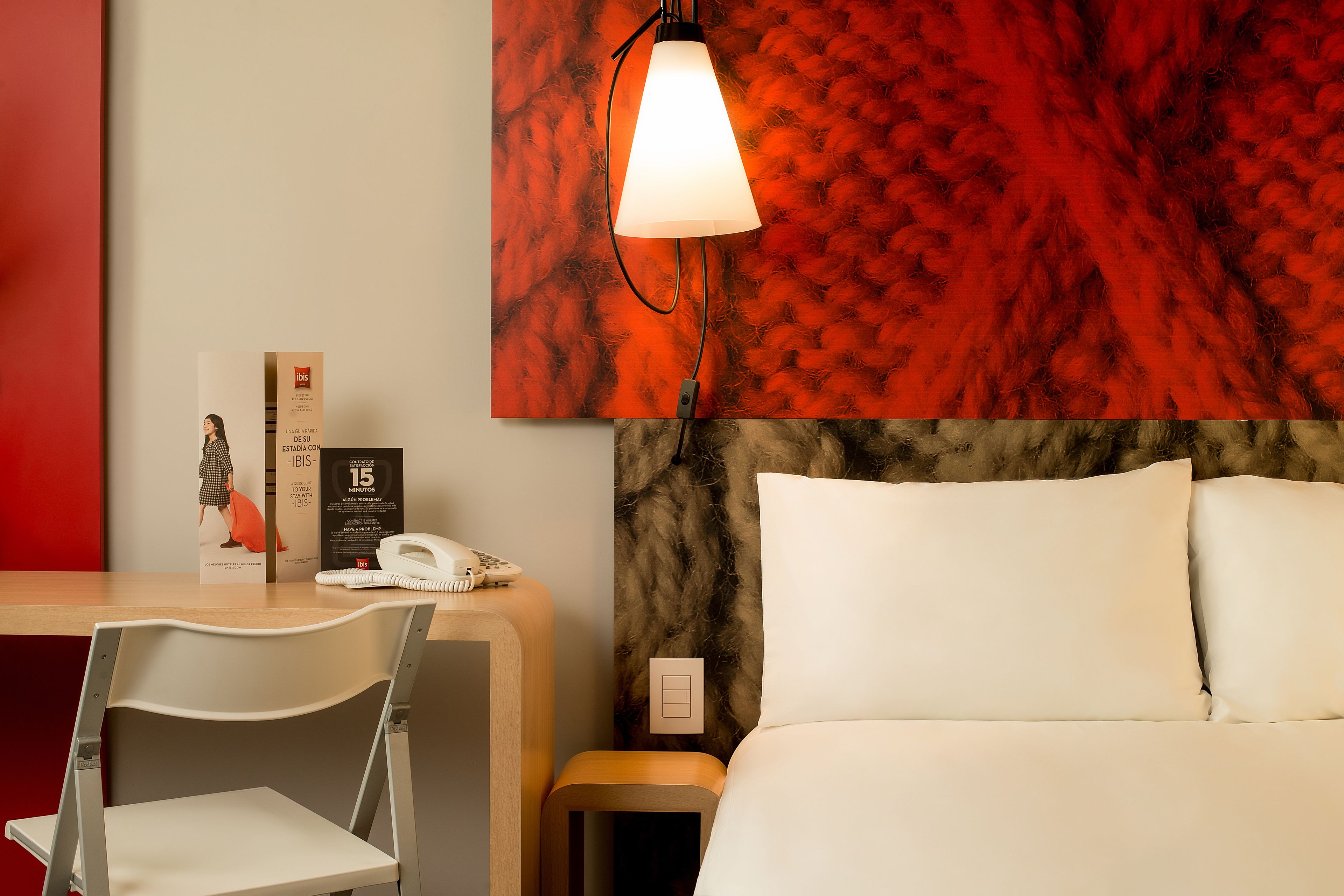 A hotel room with a bed, a nightstand, a chair, a desk, a lamp, and a red wall with a textured pattern.