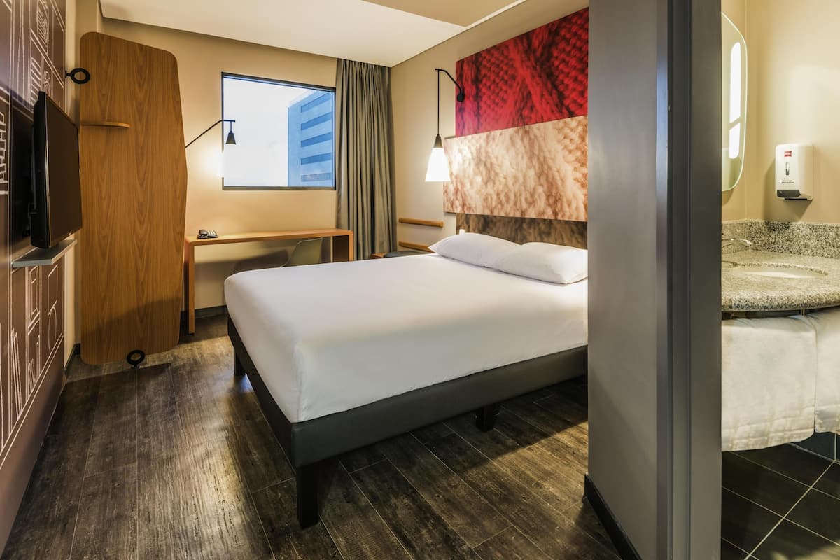 A hotel room with a bed, desk, TV, and a bathroom with a sink and mirror.