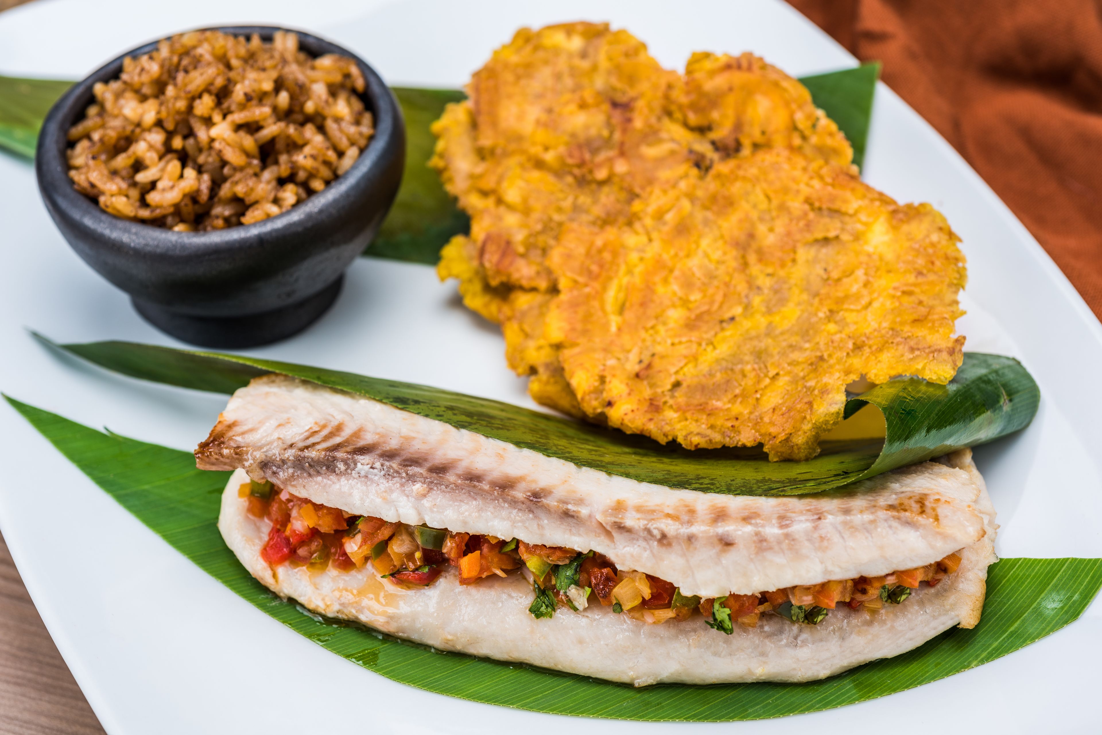 A plate with a fish sandwich, fried plantains, and a bowl of rice.