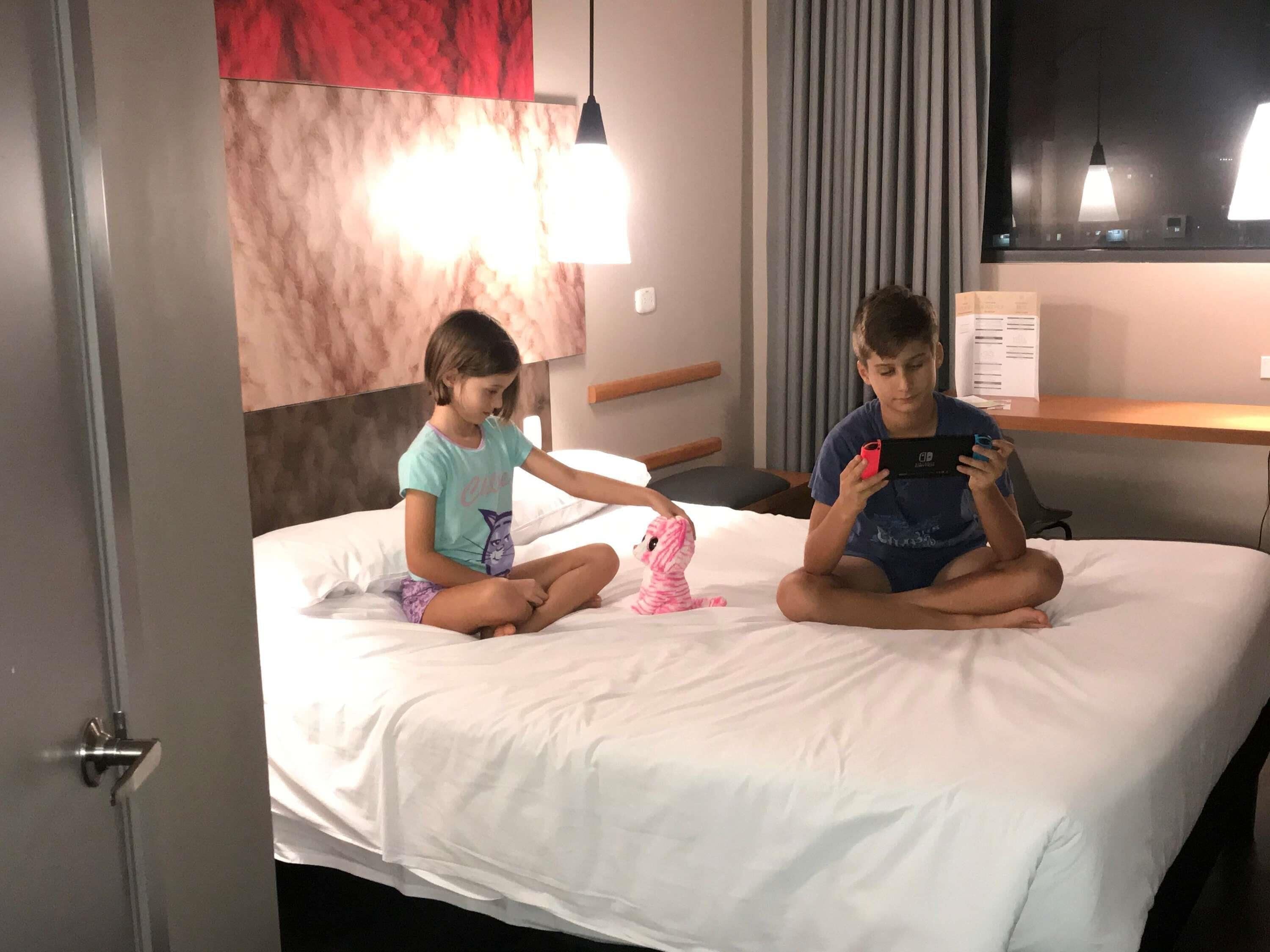 Deux enfants sont assis sur un lit dans une chambre d’hôtel. Le mur est rouge et il y a une fenêtre avec des rideaux.