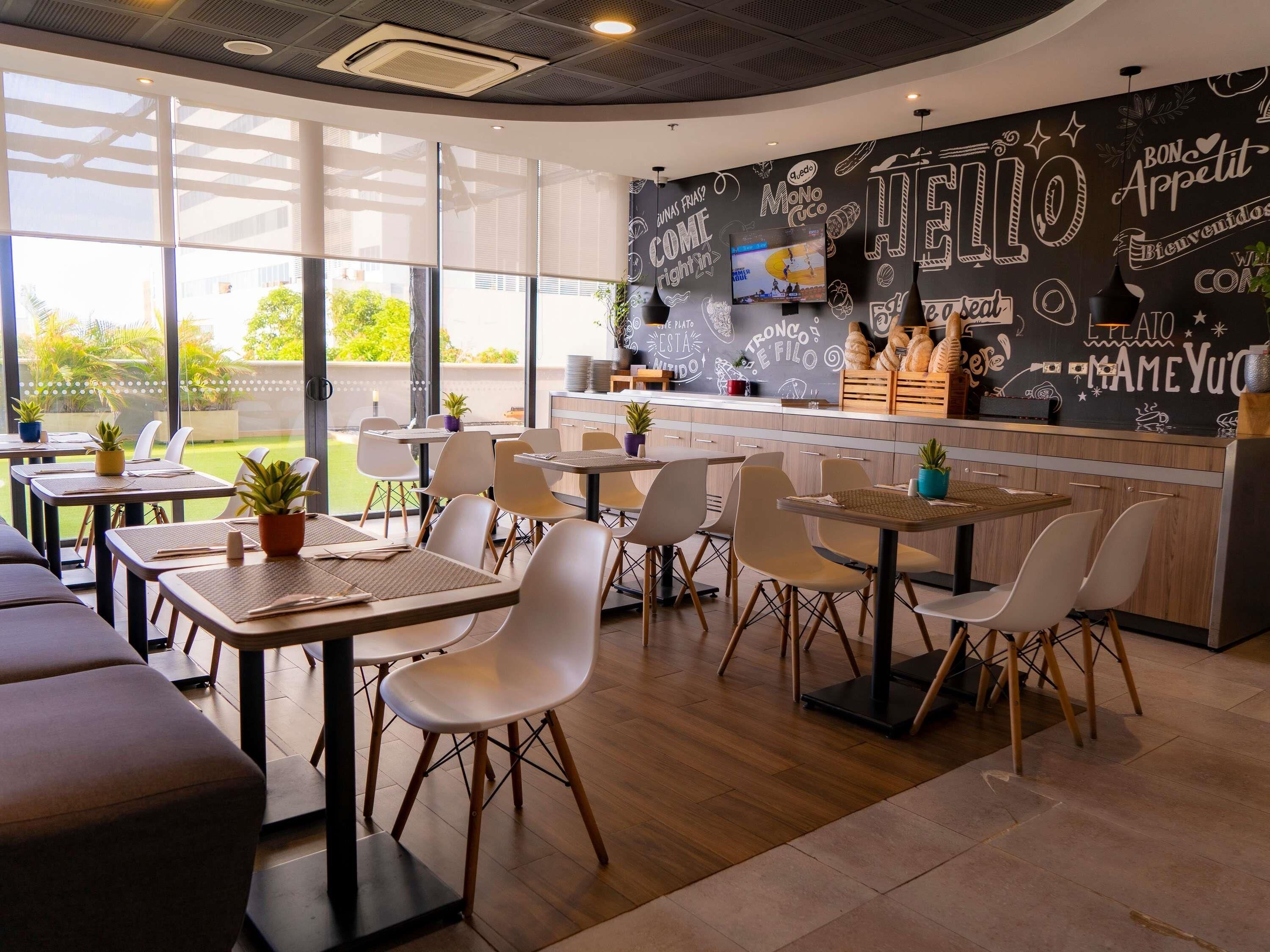 Un café moderne avec un mur noir où sont dessinés divers gribouillages et où figure du texte, un comptoir avec une machine à café et des tables à l’extérieur agrémentées de plantes en pot.