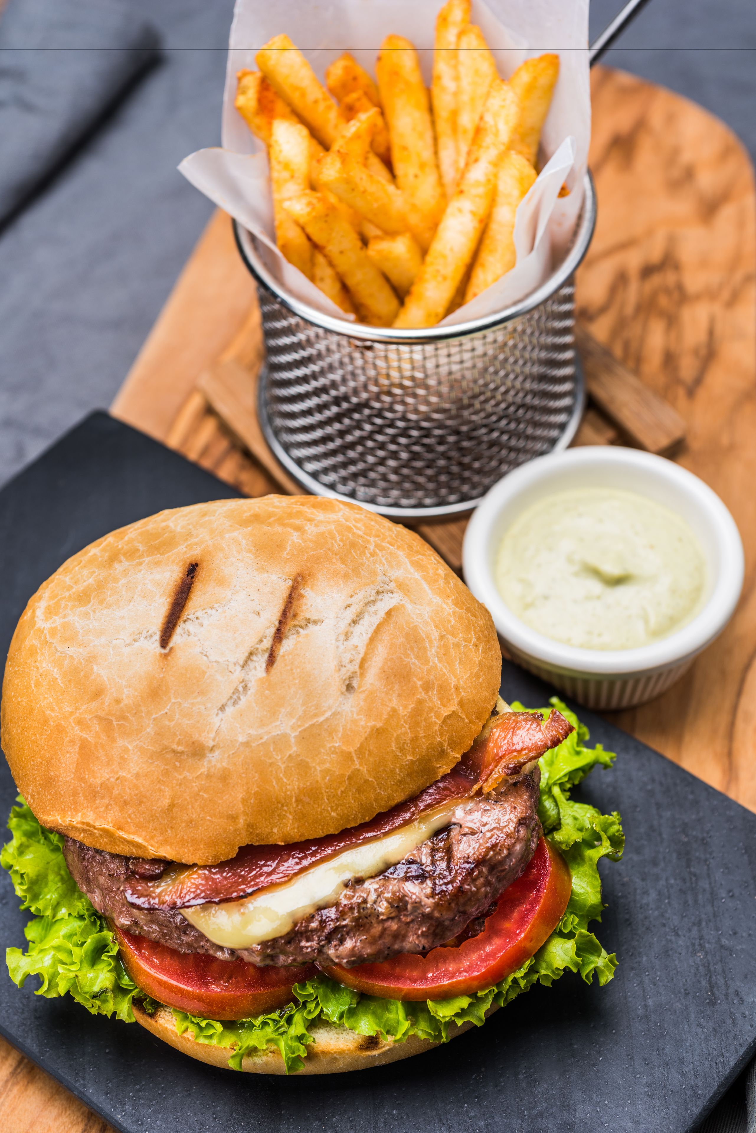 A burger with lettuce, tomato, and bacon, a basket of fries, and a small bowl of sauce on a wooden table.