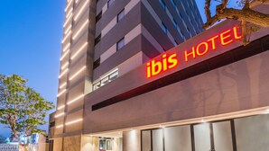 A modern hotel building with a prominent "ibis HOTEL" sign, featuring a vertical layout with multiple floors and a flat roof.