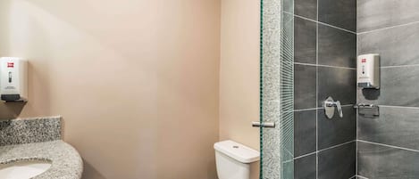Egy fürdőszoba, amelyben található egy WC, egy mosdókagyló, egy üvegfalas zuhanykabin és egy kézfertőtlenítő-adagoló.