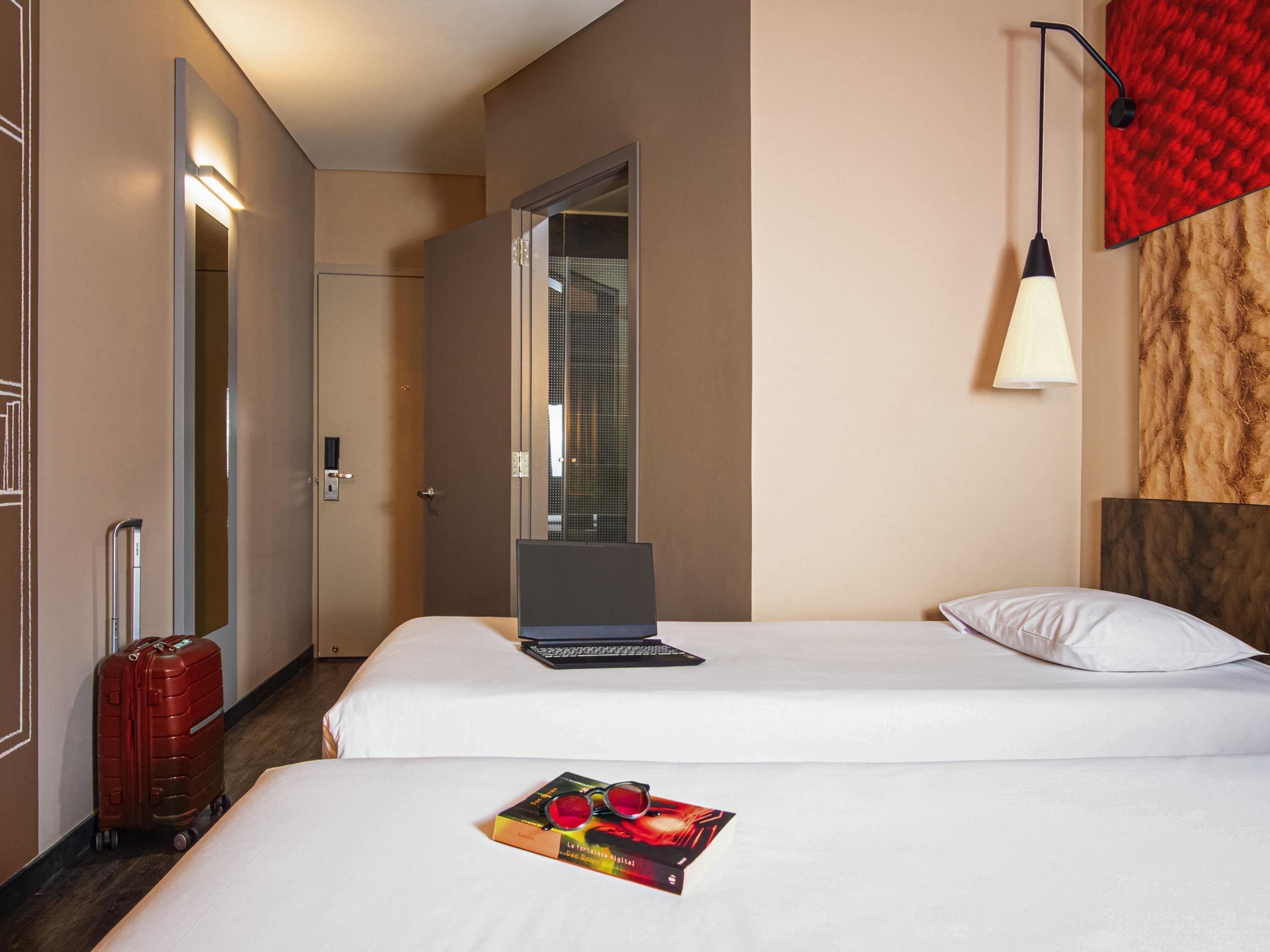 A hotel room with two beds, a laptop on one bed, a suitcase on the floor, and a mirror on the wall.