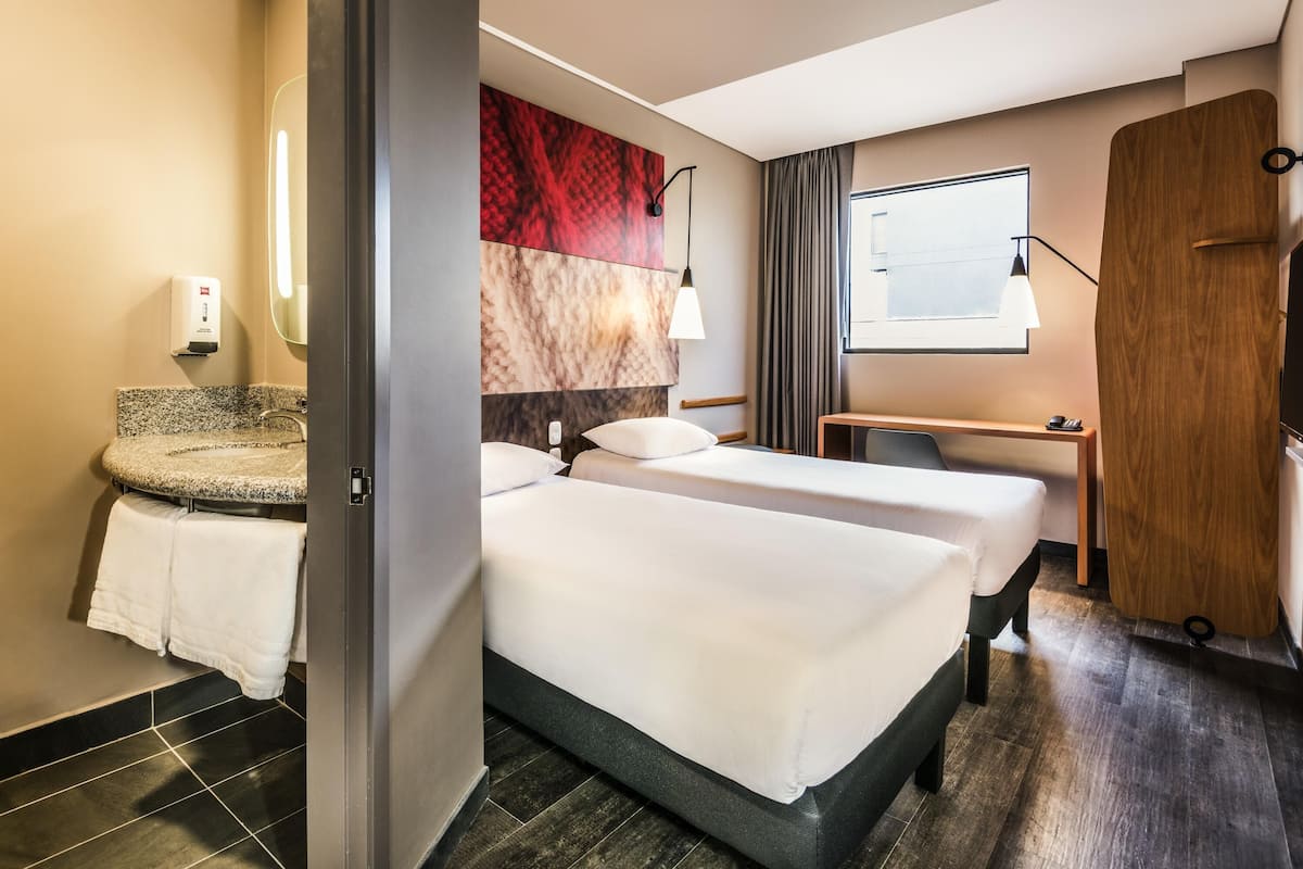 A hotel room with a bed, a desk, a chair, a bathroom with a sink, and a mirror.