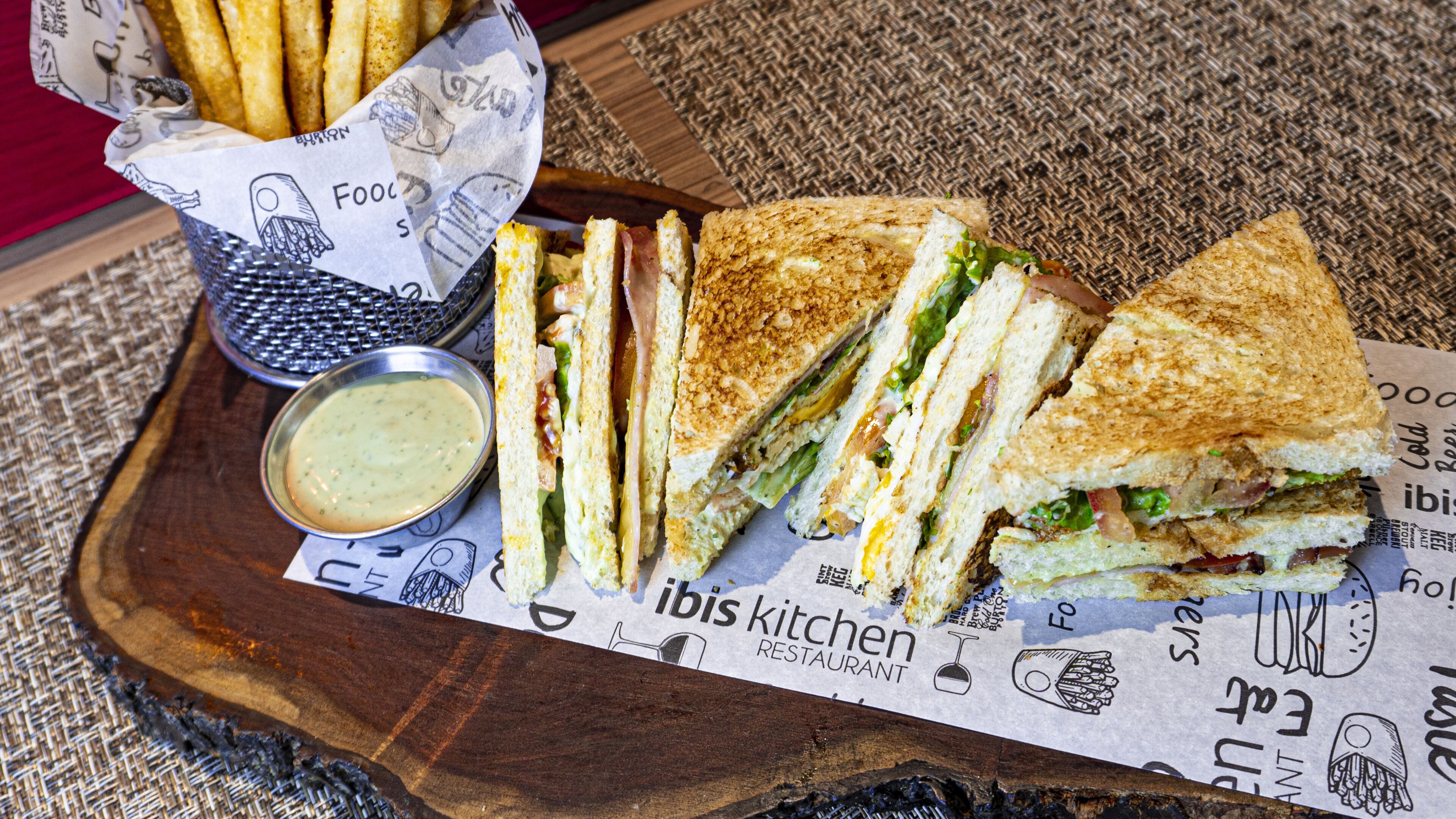 A sandwich cut in half, a small container of sauce, and a basket of fries on a wooden board with a paper underneath.