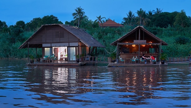 Two wooden structures with open porches, situated on a body of water with a backdrop of dense greenery and palm trees.