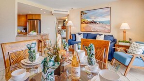 Een moderne woonkamer met een eethoek, een keuken op de achtergrond en een schilderij met een strandthema aan de muur.