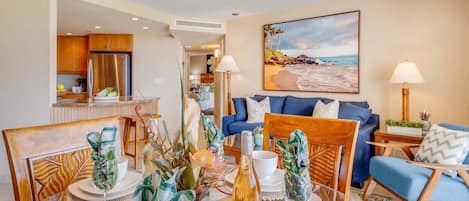 Uma sala de estar moderna com área de jantar, cozinha ao fundo e um quadro com tema praiano na parede.
