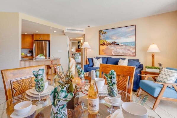 A modern living room with a dining area, a kitchen in the background, and a beach-themed painting on the wall.