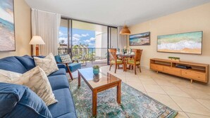 Een ruime woonkamer met een blauwe bank, een salontafel en een televisie aan de muur. Er is een balkon met uitzicht op de oceaan.