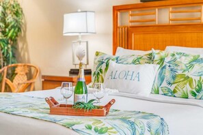 A neatly made bed with a tropical-themed bedspread, a wooden tray with a bottle and glasses, and a pillow with "ALOHA" on it.