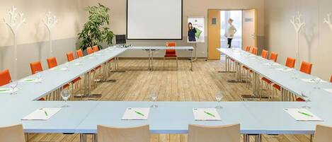Un salón de conferencias con una larga mesa, sillas anaranjadas y una pantalla de proyector.