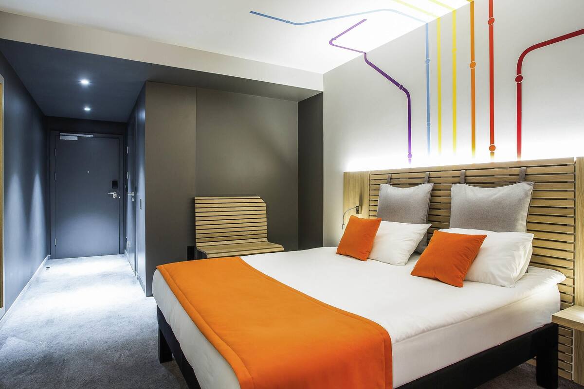 A modern hotel room with a large bed, a wooden headboard, and a bright ceiling with colorful lines.