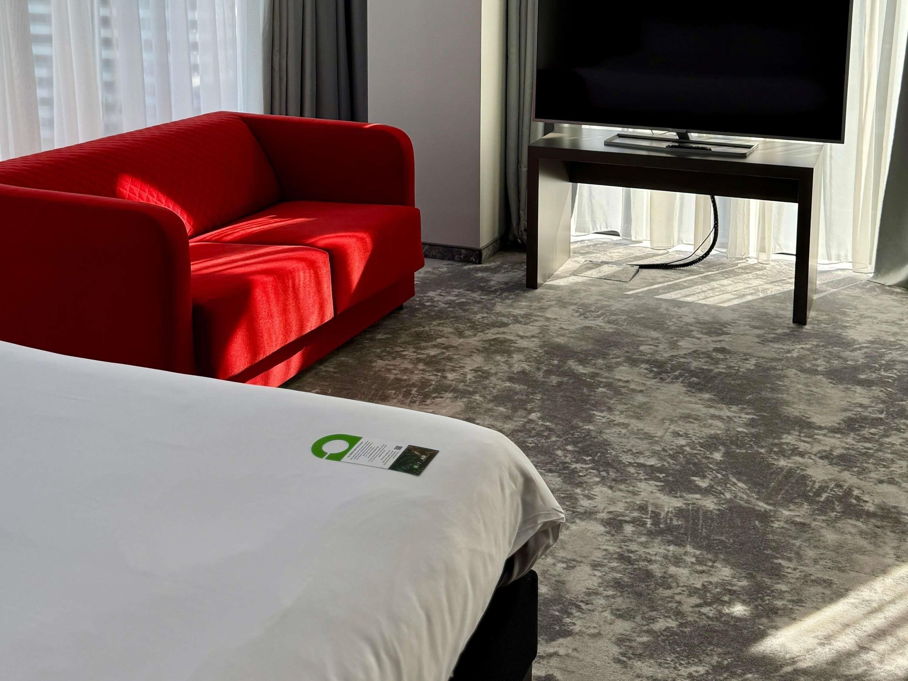 A hotel room with a bed, a red armchair, a television on a stand, and a window with curtains.