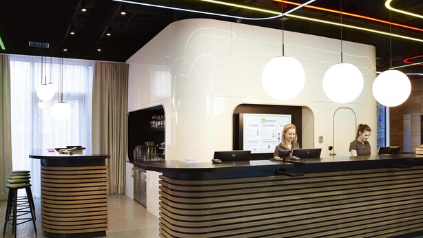 A modern reception desk with two staff members, a bar area, and a large screen displaying information. - ibis Styles Warszawa City (Warsaw)