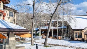 A snow-covered courtyard with modern buildings, a wooden deck, and a clear sky. - 1107 Trappeur's Lodge, Trappeur's Crossing Apartment 1 (Steamboat Springs)