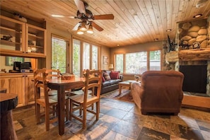 A cozy cabin interior with a dining area, sofa, fireplace, and wooden ceiling.