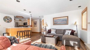 A modern living room with a sofa, ottoman, and coffee table. A kitchen is visible in the background with a bar stool. - 1112 Trappeur's Lodge, Trappeur's Crossing Apartment 4 (Steamboat Springs)