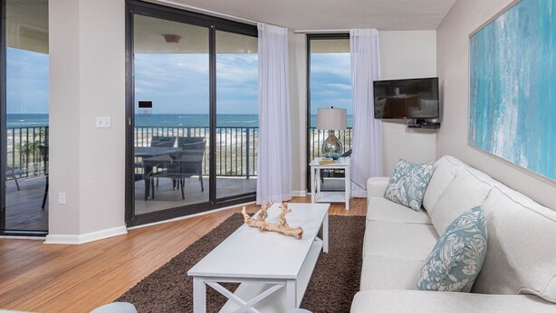A living room with a view of the beach, a white sofa, a coffee table, and a TV on the wall.