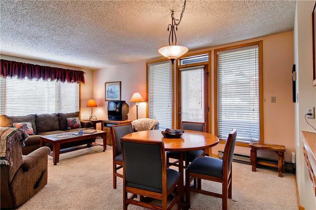 A cozy living room with a sofa, coffee table, and dining table with chairs. There is a television, lamp, and windows with blinds.