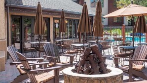 An outdoor patio with wooden chairs and tables, surrounded by umbrellas, and a stone fire pit in the center.