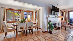 Un salon spacieux avec une cheminée, un coin repas avec une table et des chaises, et de grandes fenêtres avec des rideaux.