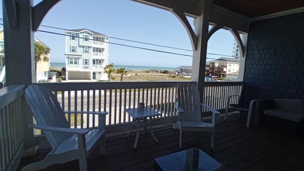 A covered porch with white wooden chairs and a table, overlooking a beach and buildings. - Fabulous views  - right across the street from the beach! (Carolina Beach)
