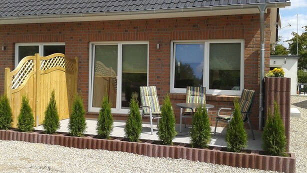 A two-story brick house with a grey tiled roof, a wooden gate, and a patio area with four chairs and small potted plants. - Apartment Berger - Apartment (Timmendorfer Strand)