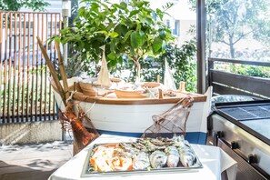 A grill with seafood on a table, a boat with a net, and a tree in the background. - BARDOLINO Fine.Food.Hotel (Nuremberg)