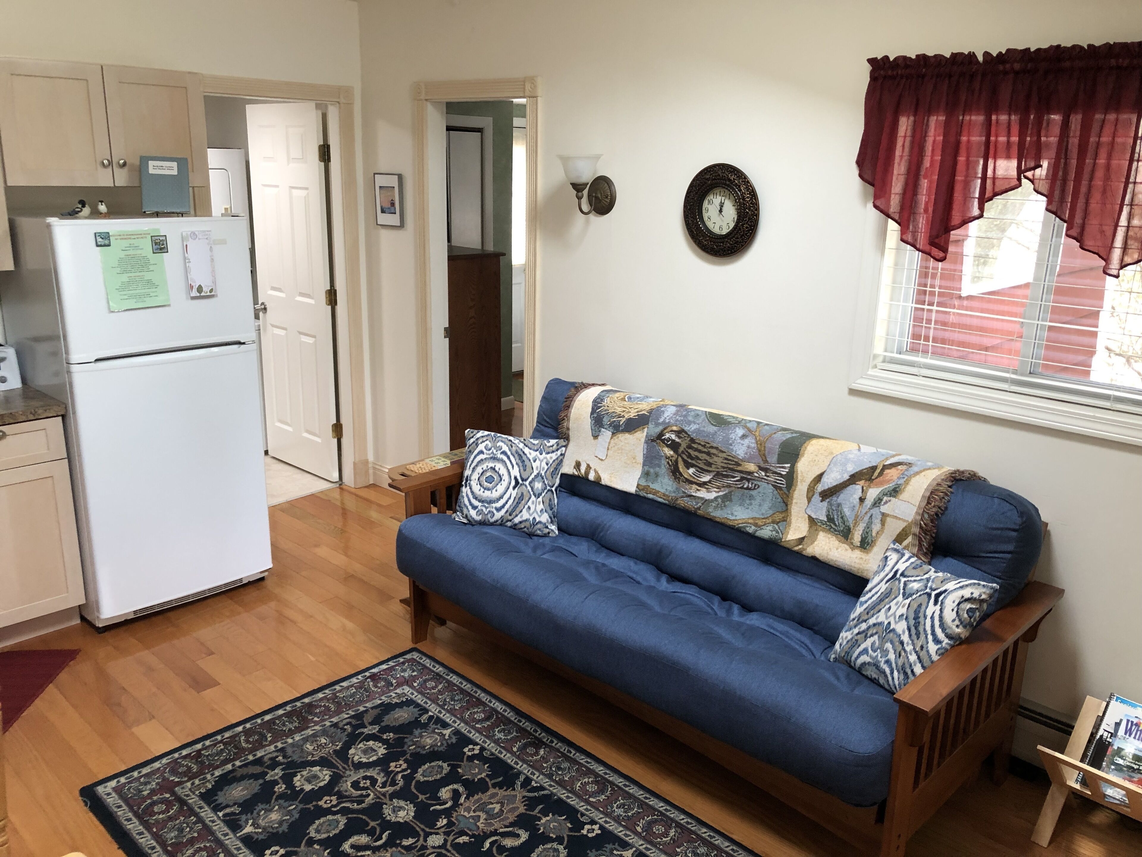 A living room with a blue sofa, a patterned rug, a wall clock, and a window with a red curtain.