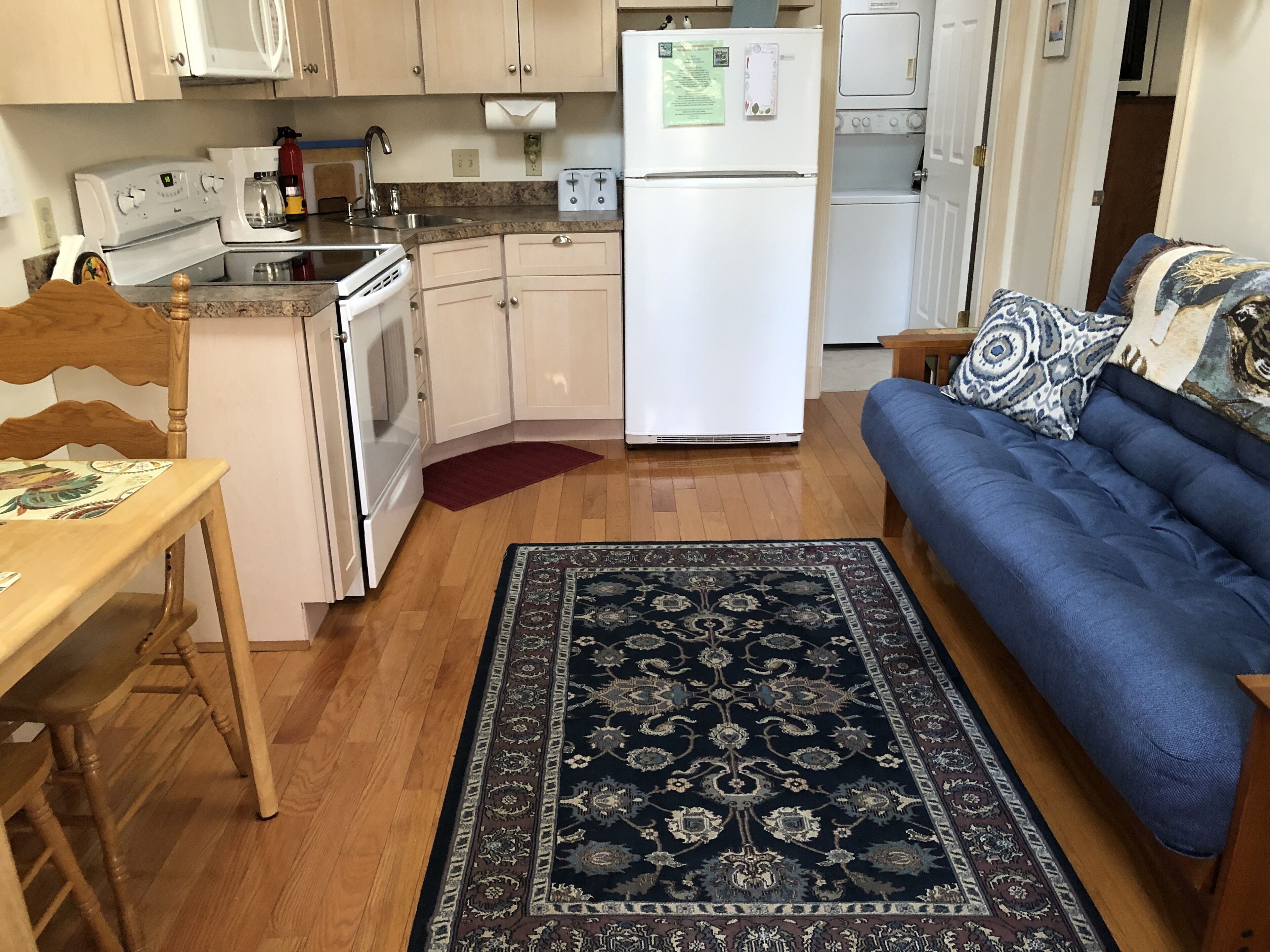 A compact kitchen with a stove, sink, and refrigerator. There is a blue sofa and a patterned rug in the living area.