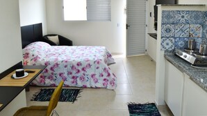 A compact room with a bed, a small table with a cup, a chair, a window, and a kitchenette with a stove. - Studio Ana Laura (São Paulo)