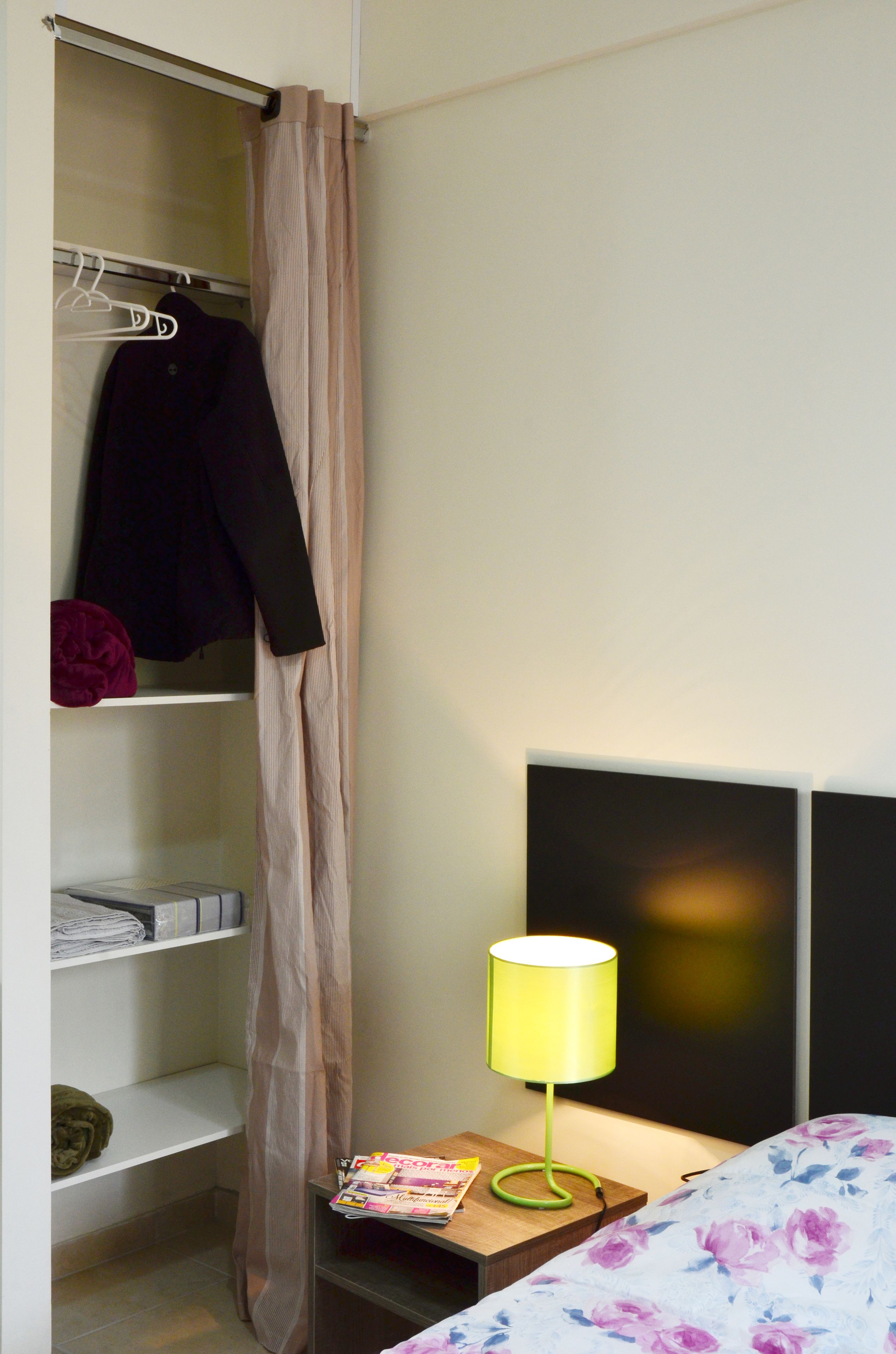A bedroom with a bed, a nightstand with a lamp, a closet with hanging clothes, and a shelf with folded items.
