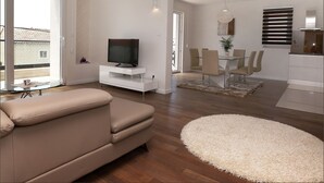 Una sala de estar moderna con un sofá, un televisor sobre un mueble blanco, una zona de comedor con sillas y una mesa, y un ventanal con persianas.