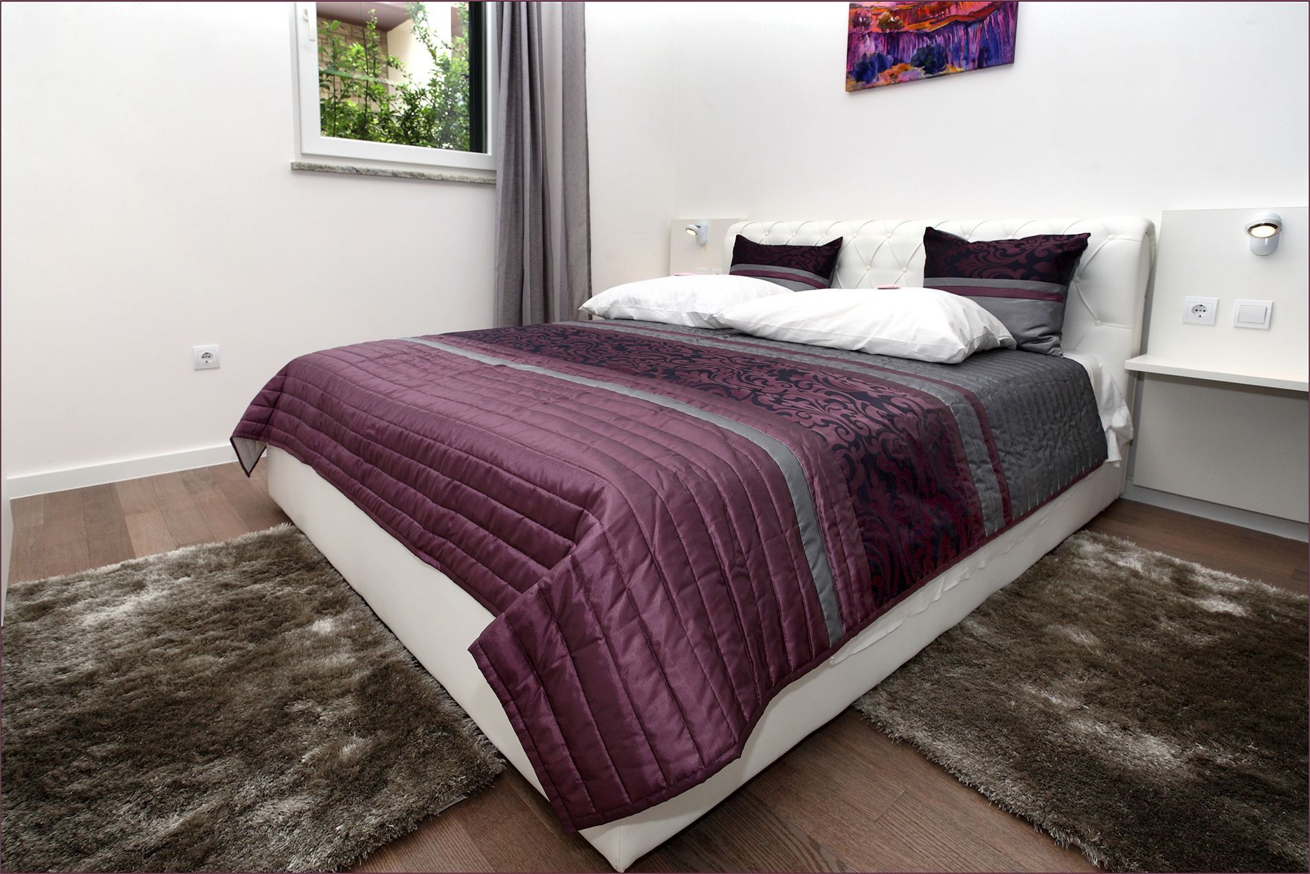 Ein ordentlich bezogenes Bett mit einer violett-grauen Decke, einem flauschigen Teppich, einem Fenster mit Blick auf Grünflächen und einem Bild an der Wand.