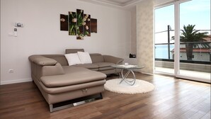 Una sala moderna con un sofá seccional color beige, una mesa de centro de vidrio y un gran ventanal corredizo que da a un balcón con vista al mar.