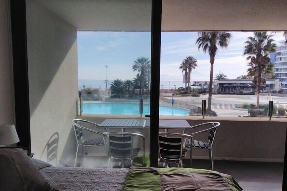 A hotel room with a bed, a table, and chairs. There is a view of a pool and palm trees outside.