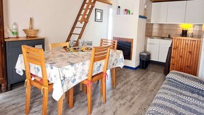 A dining area with a table set for four, wooden chairs, a wooden ladder, a kitchen with white cabinets, and a painting on the wall. - 2 Room Mais., Pool, BBQ, Near the Sea (Bretignolles-sur-Mer)