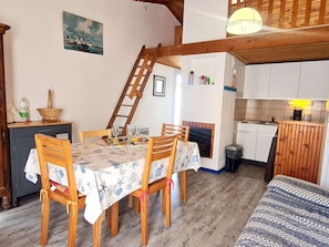 Un comedor con una mesa dispuesta para cuatro personas, sillas de madera, una escalera de madera, una cocina con armarios blancos y un cuadro en la pared.