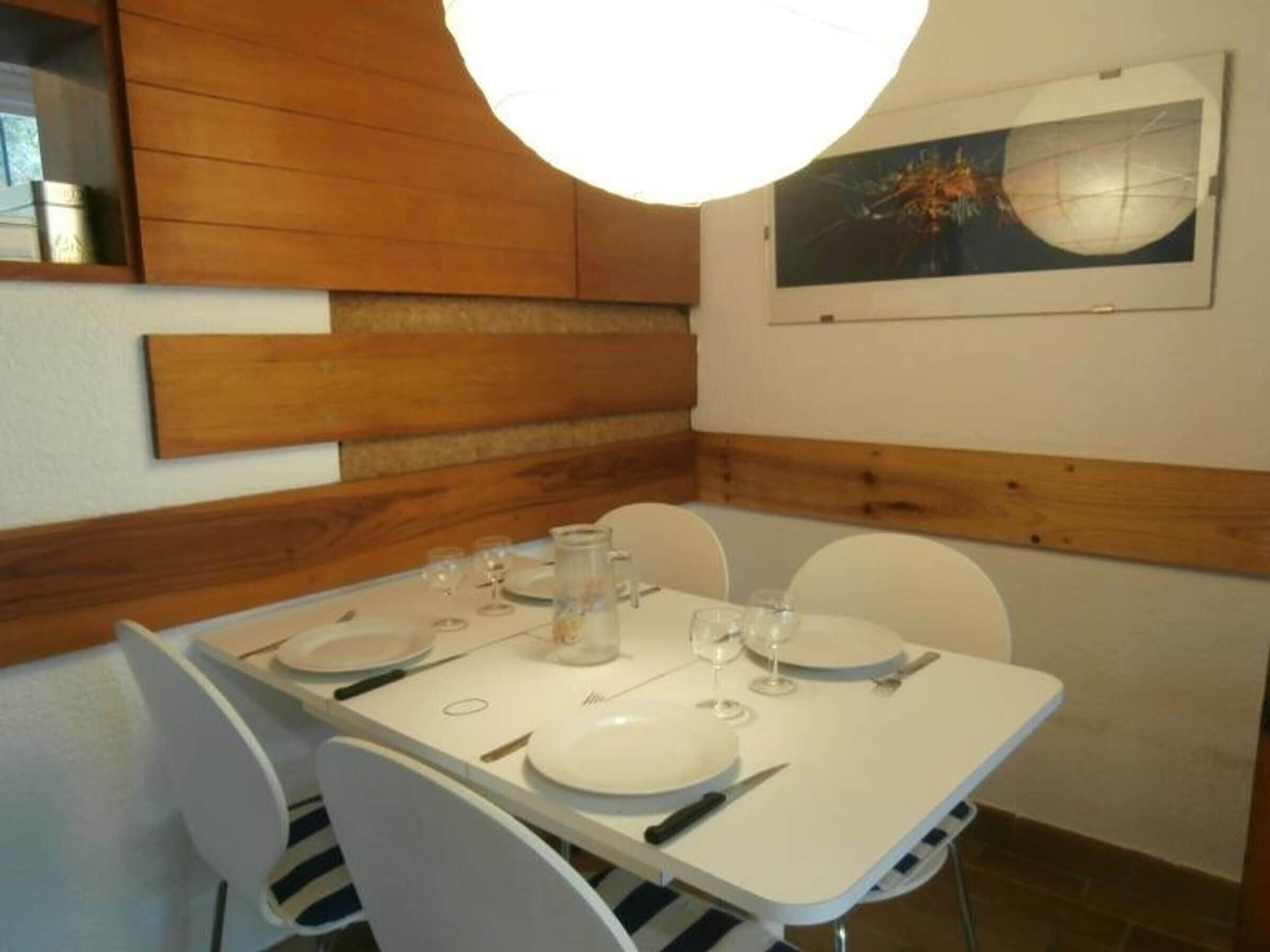 Un comedor con una mesa dispuesta para cuatro personas, sillas blancas y un cuadro en la pared.